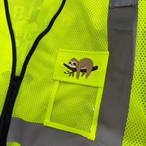 Op de afbeelding: Een neongeel veiligheidsvest met hoge zichtbaarheid van mesh-stof met zwarte randen en reflecterende zilveren verticale strepen. Een klein rechthoekig zakje aan de voorzijde toont een grafische illustratie van een bruine luiaard op een tak.