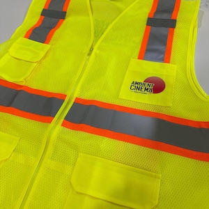 Op de afbeelding: Een neongeel veiligheidsvest met hoge zichtbaarheid van mesh-stof. Het is voorzien van verticale en horizontale zilveren reflecterende strips met oranje randen. De linkerborstzak toont een logo met de tekst Ambient Cinema Solutions.