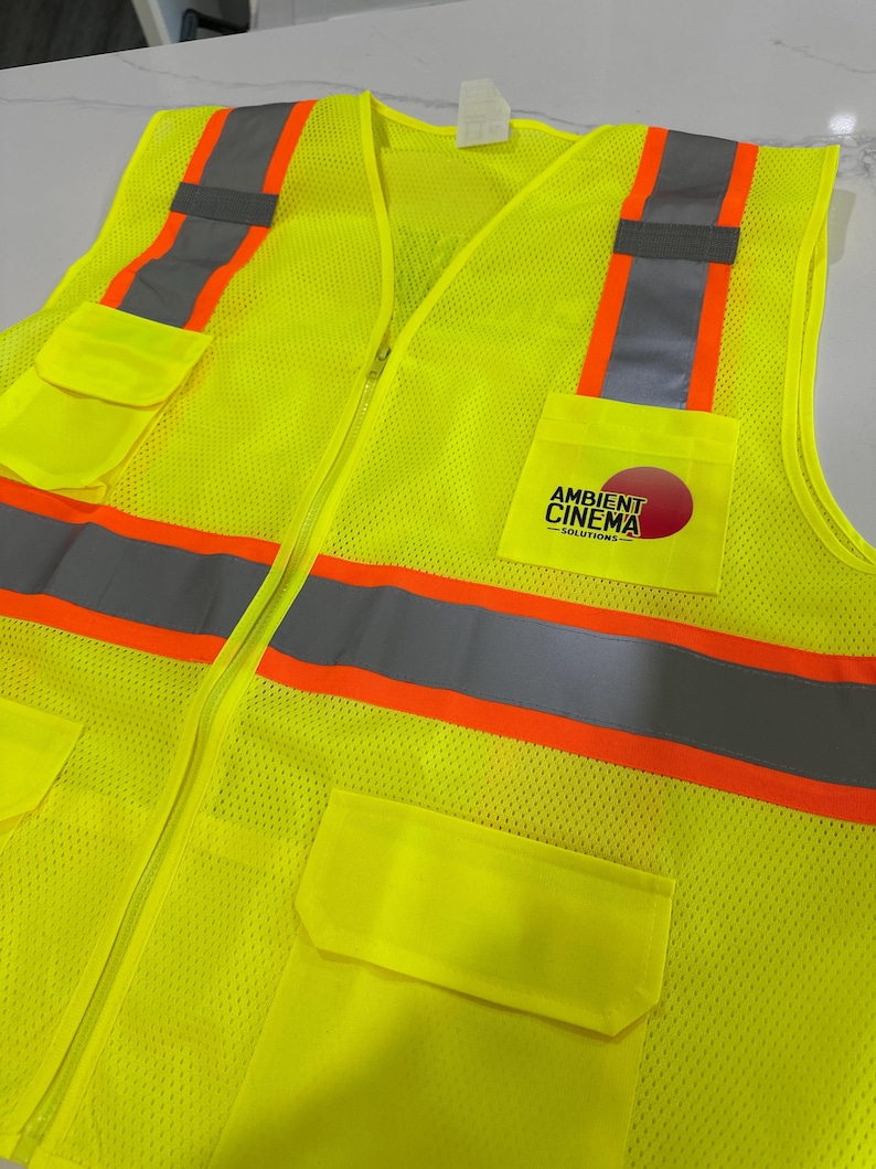 Op de afbeelding: Een neongeel veiligheidsvest met hoge zichtbaarheid van mesh-stof. Het is voorzien van verticale en horizontale zilveren reflecterende strips met oranje randen. De linkerborstzak toont een logo met de tekst Ambient Cinema Solutions.
