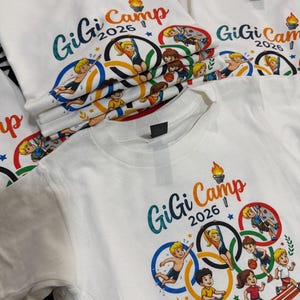 May include: White t-shirts with a colorful graphic design featuring the text "GiGi Camp 2026" and illustrations of children participating in various sports, including swimming, gymnastics, and running. The design incorporates Olympic rings.