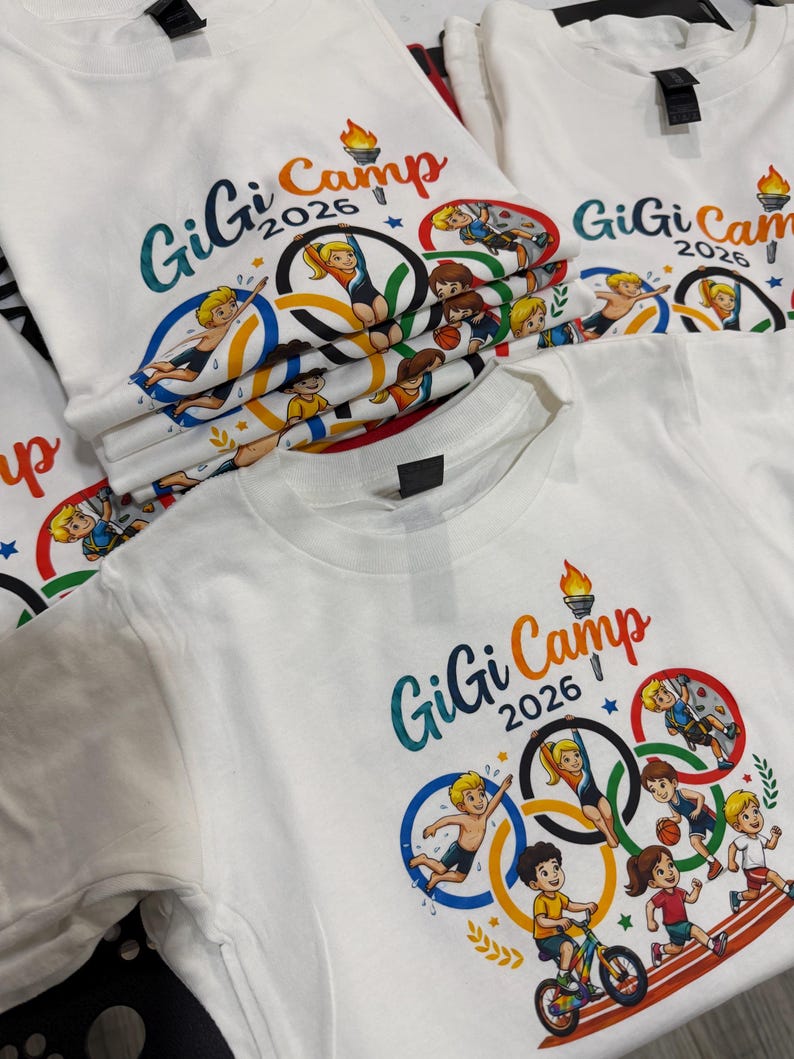 May include: White t-shirts with a colorful graphic design featuring the text "GiGi Camp 2026" and illustrations of children participating in various sports, including swimming, gymnastics, and running. The design incorporates Olympic rings.
