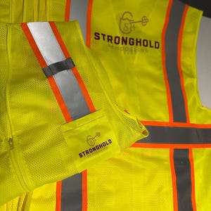 Op de afbeelding: Een neon-geel veiligheidsvest met hoge zichtbaarheid en reflecterende zilveren en oranje verticale strepen. Op de achterkant en een klein voorvak staat een logo met een sleutelpictogram en de tekst Stronghold Remodeling in donkerbruin.