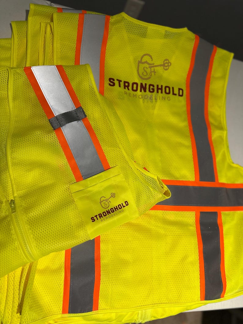 Op de afbeelding: Een neon-geel veiligheidsvest met hoge zichtbaarheid en reflecterende zilveren en oranje verticale strepen. Op de achterkant en een klein voorvak staat een logo met een sleutelpictogram en de tekst Stronghold Remodeling in donkerbruin.
