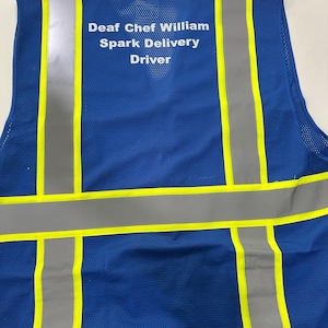 Op de afbeelding: Een blauw veiligheidsvest van mesh met verticale en horizontale reflecterende zilveren strepen, afgezet met neongele randen. Op de achterkant staat de witte tekst: Deaf Chef William, Spark Delivery, Driver.