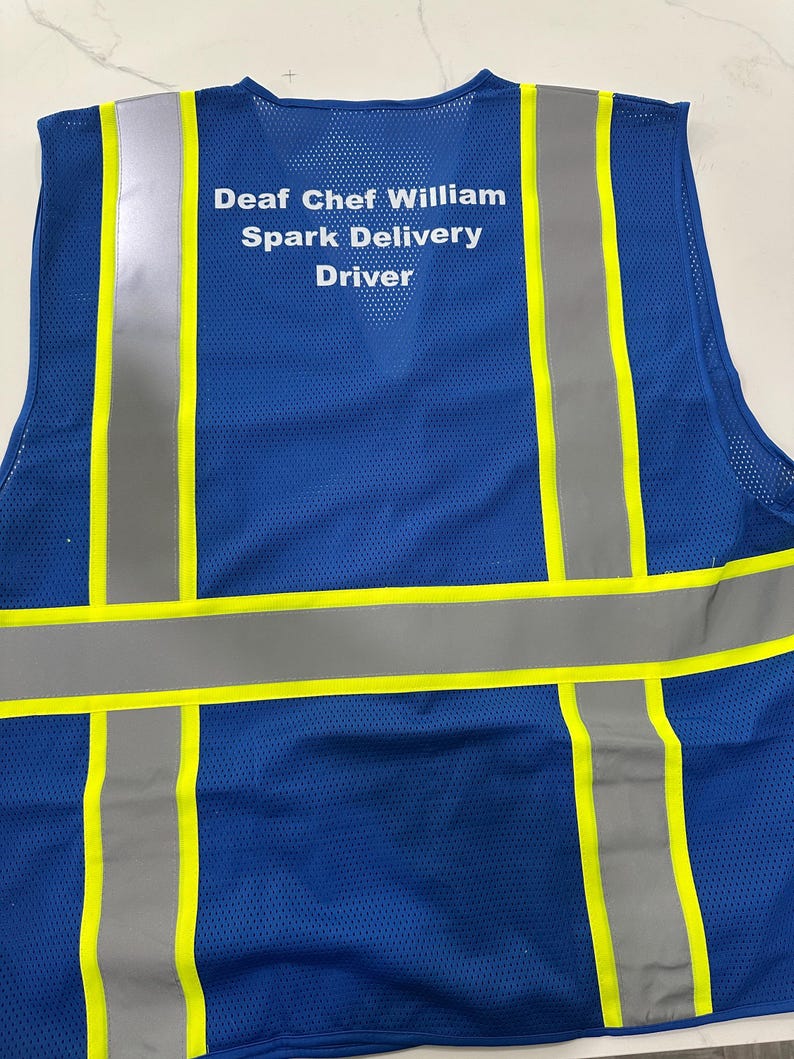 Op de afbeelding: Een blauw veiligheidsvest van mesh met verticale en horizontale reflecterende zilveren strepen, afgezet met neongele randen. Op de achterkant staat de witte tekst: Deaf Chef William, Spark Delivery, Driver.