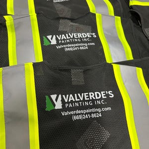 Op de afbeelding: Zwarte veiligheidsvesten van mesh met neongele reflecterende verticale strepen. Op de achterkant van elk vest staat het wit-groene logo van Valverde's Painting Inc., de website Valverdespainting.com en het telefoonnummer (669)241-8624.