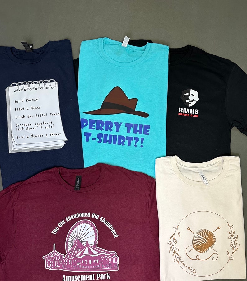 May include: A collection of folded t-shirts in various colors. One navy blue shirt has a notepad graphic with a list. A turquoise shirt features a brown hat graphic and text. Other shirts are black, burgundy, and cream-colored.