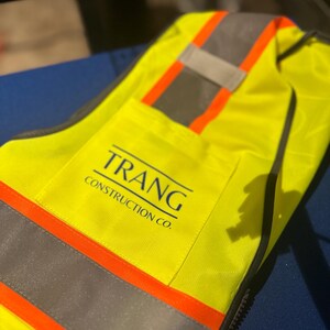 Op de afbeelding: Een neongeel veiligheidsvest met horizontale zilveren reflecterende strips omzoomd door feloranje banden. Op de borst staat de tekst TRANG CONSTRUCTION CO. in donkerblauwe letters met een horizontale lijn erboven.