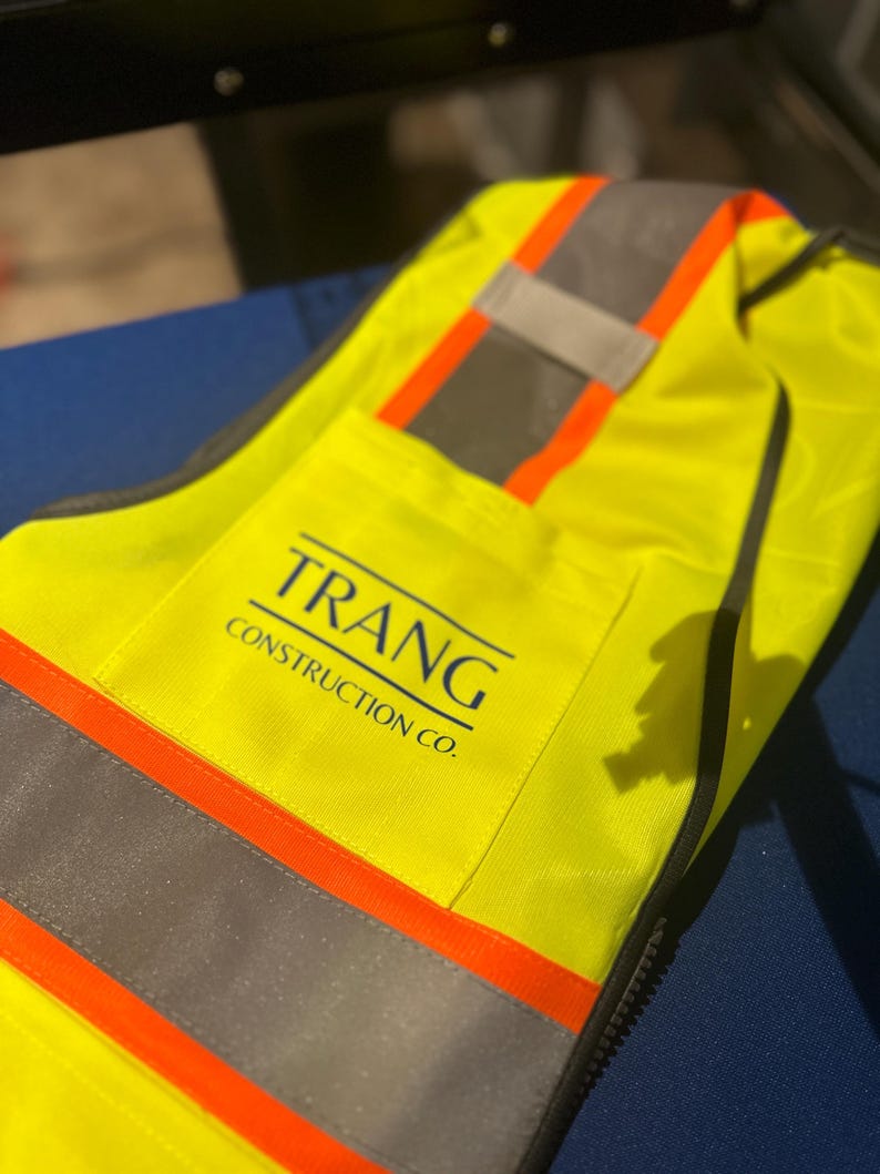 Op de afbeelding: Een neongeel veiligheidsvest met horizontale zilveren reflecterende strips omzoomd door feloranje banden. Op de borst staat de tekst TRANG CONSTRUCTION CO. in donkerblauwe letters met een horizontale lijn erboven.