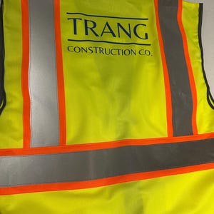 Op de afbeelding: Een neongeel veiligheidsvest met hoge zichtbaarheid, voorzien van verticale en horizontale zilveren reflecterende strips met een feloranje rand. Op de achterkant van het vest staat de donkerblauwe tekst TRANG CONSTRUCTION CO. in een vetgedrukt lettertype.