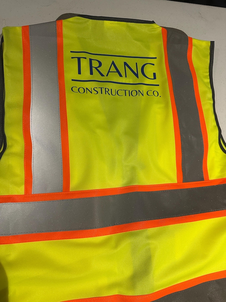 Op de afbeelding: Een neongeel veiligheidsvest met hoge zichtbaarheid, voorzien van verticale en horizontale zilveren reflecterende strips met een feloranje rand. Op de achterkant van het vest staat de donkerblauwe tekst TRANG CONSTRUCTION CO. in een vetgedrukt lettertype.
