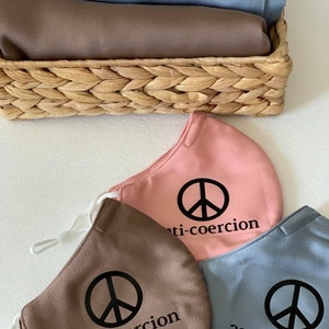May include: Three fabric face masks with a peace sign and the text "anti-coercion" printed on them. The masks are in pink, brown, and blue.
