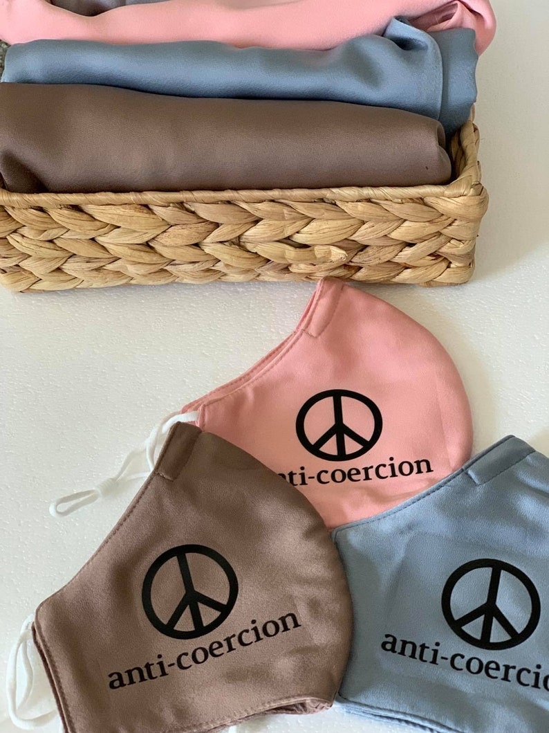 May include: Three fabric face masks with a peace sign and the text "anti-coercion" printed on them. The masks are in pink, brown, and blue.