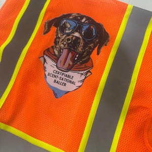 Op de afbeelding: Een feloranje veiligheidsvest met verticale zilveren reflecterende strepen en gele biezen. Een grafische print op de achterkant toont een hond met een blauwe zonnebril en een bandana met de tekst CERTIFIABLE SCENT-SATIONAL BALLER.