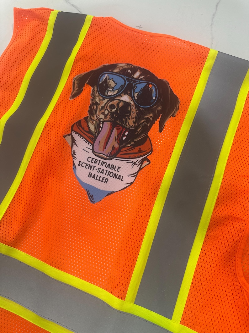 Op de afbeelding: Een feloranje veiligheidsvest met verticale zilveren reflecterende strepen en gele biezen. Een grafische print op de achterkant toont een hond met een blauwe zonnebril en een bandana met de tekst CERTIFIABLE SCENT-SATIONAL BALLER.