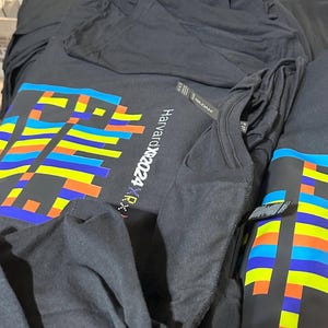 May include: Black t-shirts with a colorful pixelated design and the text "Harvardx2024 RX" are stacked on a table. The design incorporates blue, yellow, orange, and green rectangular shapes. The shirts appear to be made of a soft material.