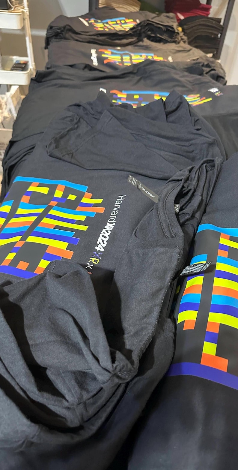 May include: Black t-shirts with a colorful pixelated design and the text "Harvardx2024 RX" are stacked on a table. The design incorporates blue, yellow, orange, and green rectangular shapes. The shirts appear to be made of a soft material.