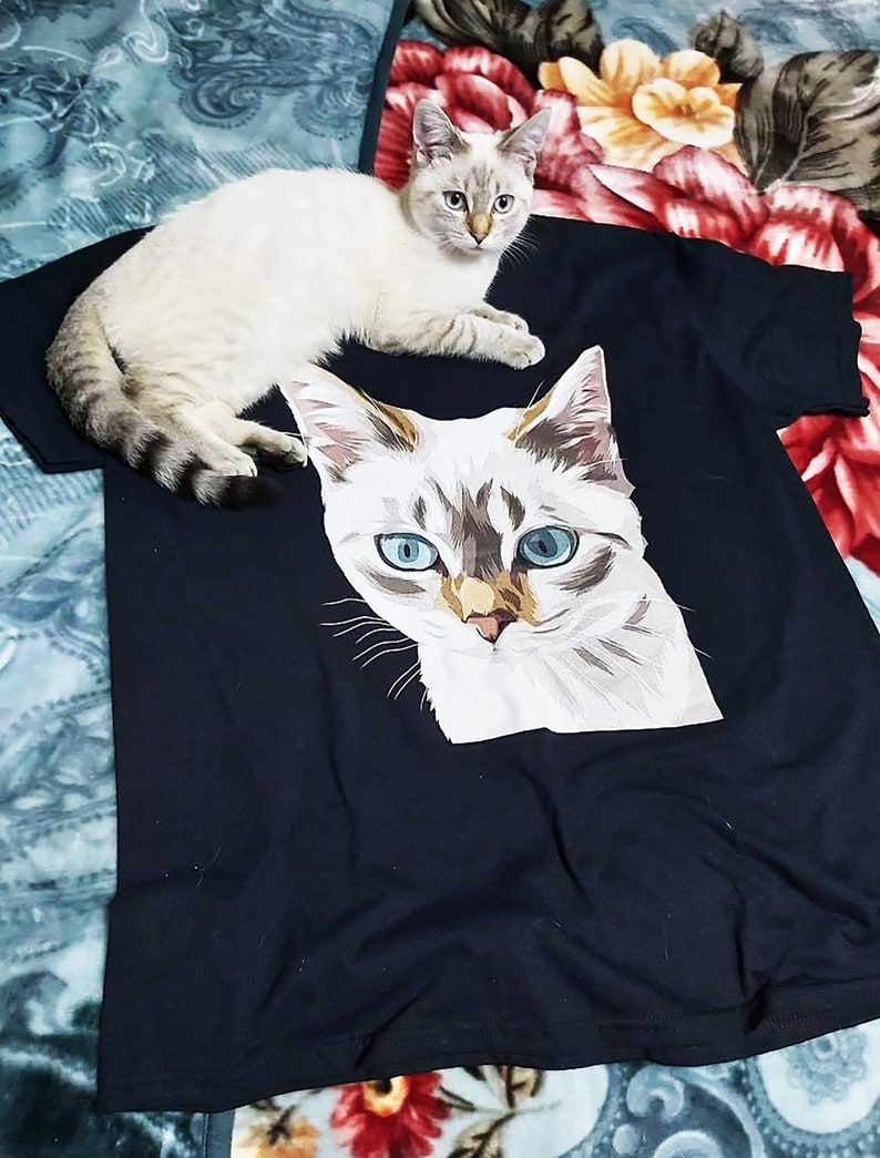 Pu&ograve; includere: Una maglietta nera con una stampa grafica di un gatto bianco. Il gatto ha gli occhi azzurri e un viso con un motivo bianco e marrone. Un gatto bianco con gli occhi azzurri &egrave; sdraiato sopra la maglietta.