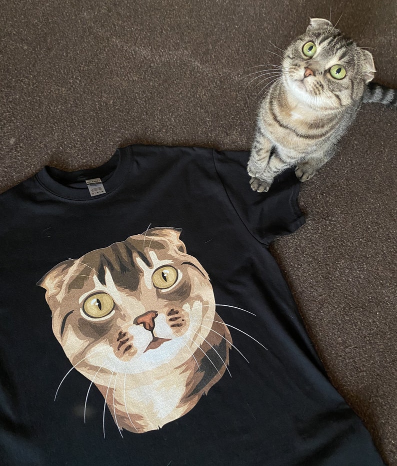 Pu&ograve; includere: Una maglietta nera con un'immagine stampata di un gatto marrone e bianco con occhi verdi. Il gatto sta guardando in alto e ha la bocca leggermente aperta.