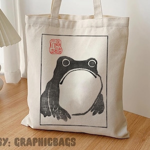May include: A cream-colored tote bag featuring a black and white frog illustration within a rectangular frame. The bag has long handles and the text "ETSY: GRAPHICBAGS" at the bottom. A small red stamp-like design is above the frog.
