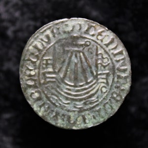 Puede incluir: Primer plano de una moneda de bronce con un diseño de barco y la inscripción "OBOLVS DEI REX ANGLIE" alrededor del borde.