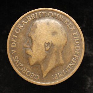 May include: A bronze coin with a profile of a man and the inscription "GEORGIVS V DEI GRA:BRITT:OMN:REX FID:DEF:IND:IMP." The coin is circular and shows intricate detailing.