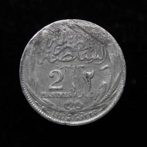 May include: A silver coin with Arabic script and the number "2" prominently displayed. The coin also features the text "PIASTRES" and the date "1917". The coin is circular and has a worn appearance, suggesting it is an antique or vintage item.
