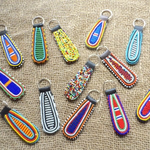 May include: A selection of beaded keyrings in diverse colours, such as blue, yellow, red, and green. Each keyring has a unique pattern and design, with a silver ring for attaching keys. The keyrings are displayed on a textured, tan surface.