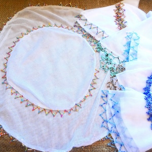 May include: White fabric items with colourful beaded trim. The image shows a large, round piece with a beaded edge, and several smaller, square pieces with blue and colourful beaded accents. The items are displayed on a textured, brown surface.