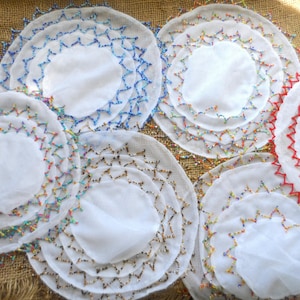 May include: A selection of white, circular doilies with detailed beaded and stitched edges. The doilies display a variety of colours, including blue, red, and multicoloured beads, giving a decorative, handcrafted look. The doilies are arranged on a textured, brown surface.