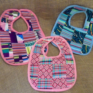 May include: Three baby bibs made of colourful patterned fabric. The bibs are pink, blue, and pink with a white trim. The bibs are made of a patchwork of different fabrics.