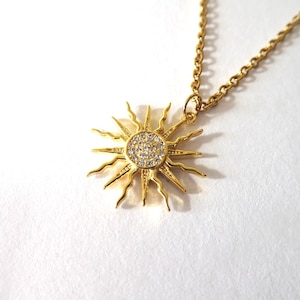 Op de afbeelding: Een gouden ketting met een zonnenhanger. De hanger is van goud en heeft een cirkel van heldere stenen in het midden. De zon heeft stralen die vanuit de cirkel naar buiten reiken.