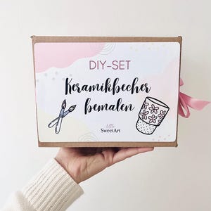 Könnte beinhalten: Eine Bastelbox mit rosa und weißem Design, mit dem Text "DIY-SET Keramikbecher bemalen" und Illustrationen von Pinseln und einer floral dekorierten Tasse. Die Box wird von einer Person gehalten.