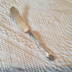 May include: A silver butter knife with an ornate handle. The handle has a floral design and a rounded end.