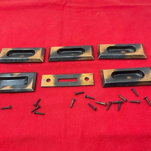 May include: A collection of six rectangular, bronze-toned drawer pulls with recessed finger grips, along with a separate, matching piece and several small screws, all arranged on a red surface. The pulls have a dark, aged appearance.