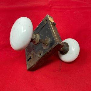 May include: Antique door hardware set featuring a black metal plate with a keyhole and two white oval porcelain doorknobs. The hardware shows signs of age and wear, with a vintage aesthetic.