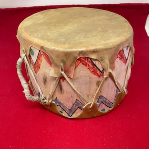 May include: A round drum with a light tan drumhead and a painted wooden body. The body is decorated with red, green, and blue geometric designs. A leather handle is attached to the side. The drum sits on a red surface.