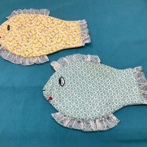 May include: Two handmade fabric fish pot holders. One is yellow with a floral pattern and the other is green with a geometric pattern. Both have white lace trim.