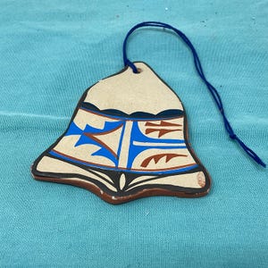 May include: A ceramic bell-shaped ornament with a beige background and a blue string. The bell features a geometric design in blue, brown, and black. The ornament has a rustic, handcrafted appearance, suitable for home decor.