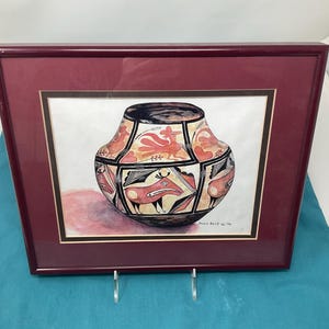 May include: A framed print of a Native American pottery jar with a red, black, and white geometric design. The jar is depicted against a white background. The print is framed in a dark red wood frame.