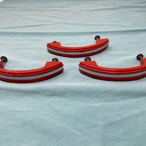 May include: Three curved drawer pulls in a bright red finish with a central light blue horizontal stripe. Each handle features two dark metal mounting screws attached to the ends, designed for installation on furniture or cabinetry doors.