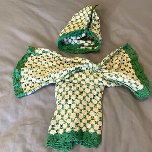 May include: A handmade, off-white and green crochet outfit, including a top and matching hat. The outfit features a checkered pattern with green trim. The hat is a rounded shape with a green trim. The outfit is displayed on a gray surface.