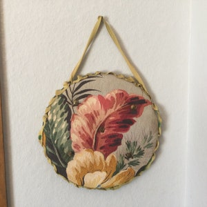 K&ouml;nnte beinhalten: Ein runder, floral gemusterter Wandbehang aus Stoff mit beigefarbenem Hintergrund und einem floralen Design in Gelb, Rot und Gr&uuml;n. Der Behang hat eine beigefarbene Stoffschlaufe zum Aufh&auml;ngen.