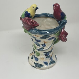 Op de afbeelding: Een keramische vaas in de vorm van een mand met een blauw en wit bloemenpatroon. Twee vogels, een gele en een rode, zitten op de rand van de vaas.