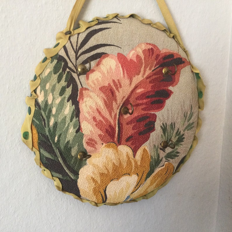 K&ouml;nnte beinhalten: Ein runder Schl&uuml;sselhalter aus Stoff mit floralem Muster. Der Stoff ist beige mit gr&uuml;nen, roten und gelben Blumen. Der Schl&uuml;sselhalter hat f&uuml;nf Metallhaken.