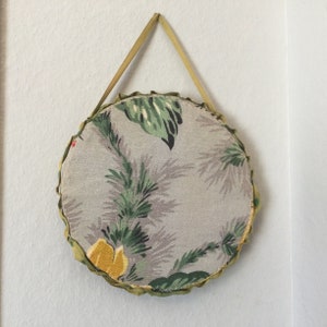 K&ouml;nnte beinhalten: Ein runder, stoffbezogener Wandbehang mit einem gr&uuml;nen und grauen Blumenmuster. Der Behang hat einen gr&uuml;nen Stoffrand und eine Schlaufe zum Aufh&auml;ngen.