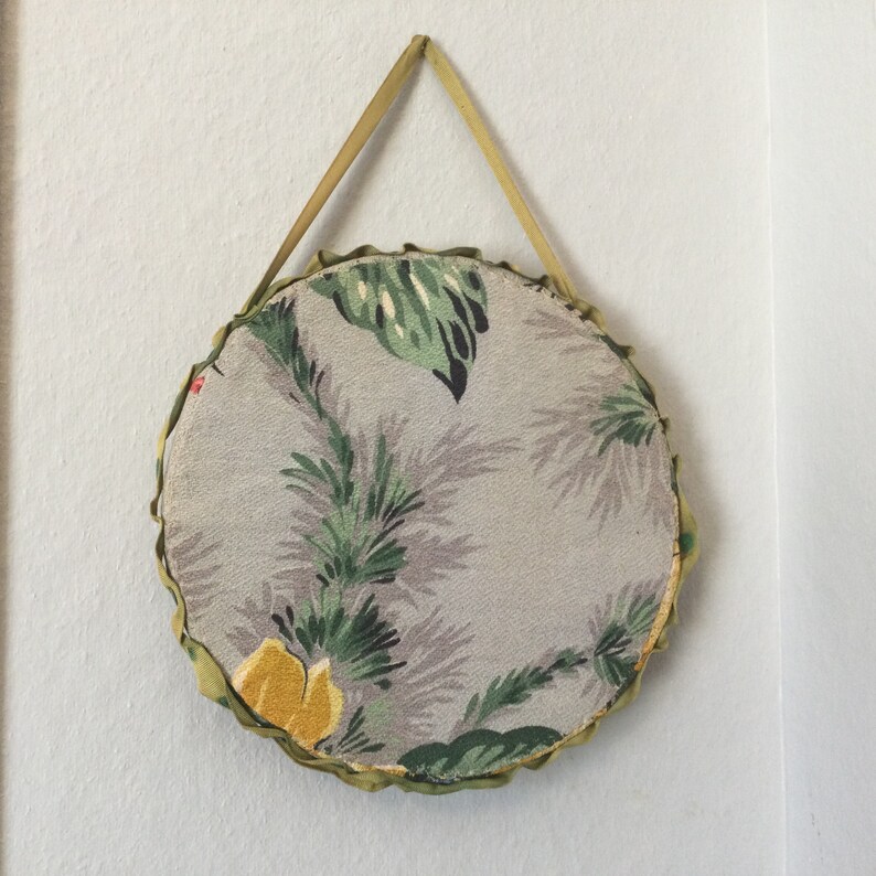 K&ouml;nnte beinhalten: Ein runder, stoffbezogener Wandbehang mit einem gr&uuml;nen und grauen Blumenmuster. Der Behang hat einen gr&uuml;nen Stoffrand und eine Schlaufe zum Aufh&auml;ngen.