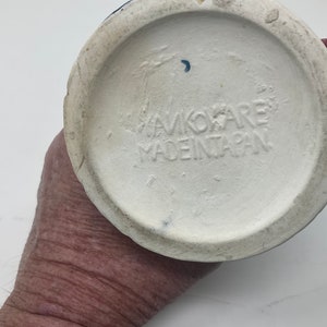 Op de afbeelding: De onderkant van een witte keramische mok met een blauw en rood ontwerp. De mok heeft de tekst "AVOKARE MADE IN JAPAN" gedrukt op de bodem.