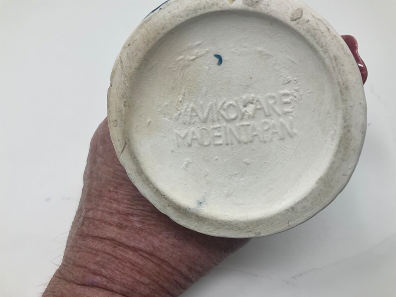 Op de afbeelding: De onderkant van een witte keramische mok met een blauw en rood ontwerp. De mok heeft de tekst "AVOKARE MADE IN JAPAN" gedrukt op de bodem.