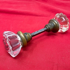 May include: Antique glass door knob with a clear, faceted design. The knob is attached to a metal rod with brass-colored hardware. The door knob is a vintage item, likely from a classic or retro style home.
